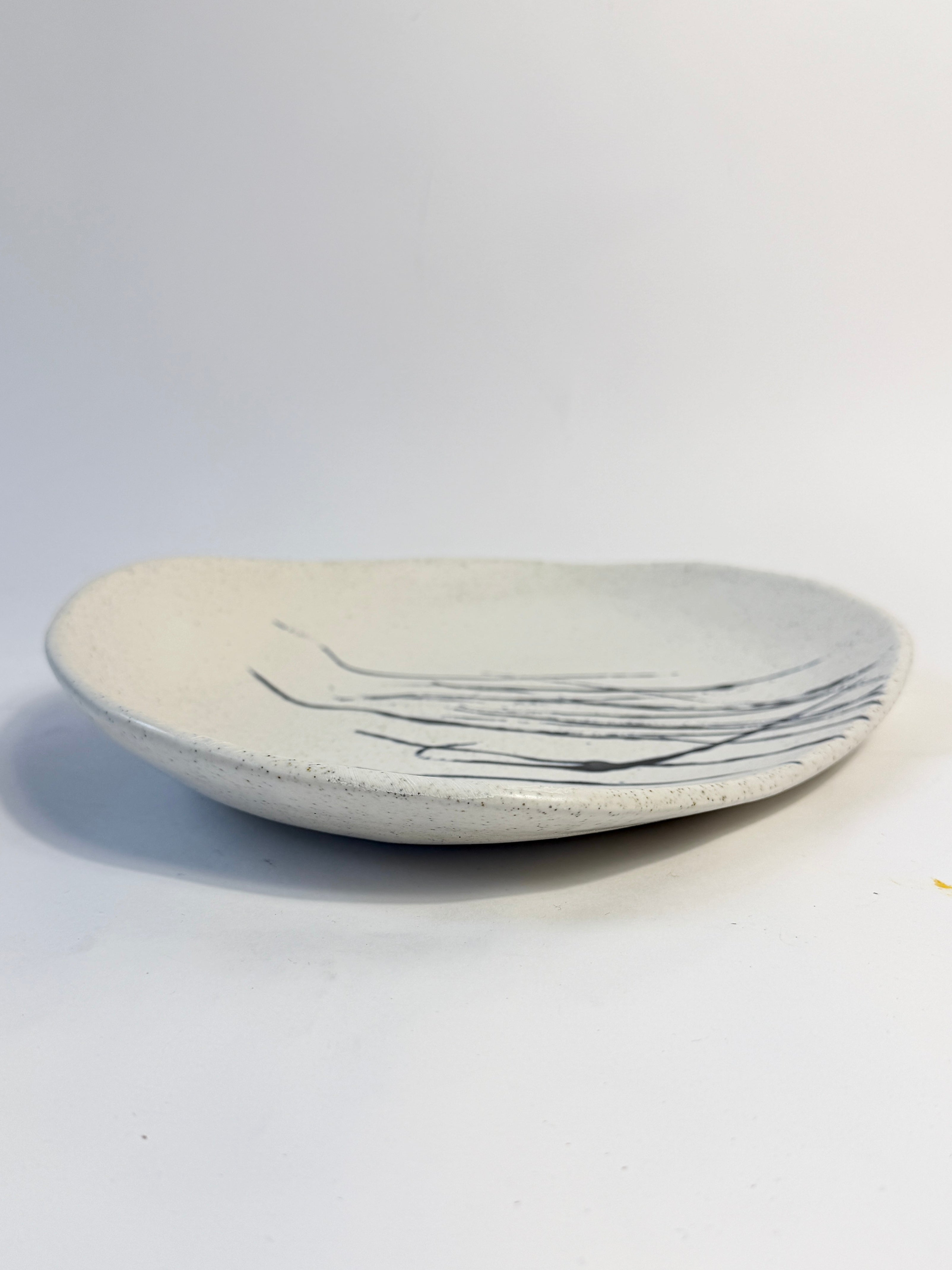 An organic shaped ceramic dish made from speckled clay with off-white glaze with dark splatter glaze decoration. 