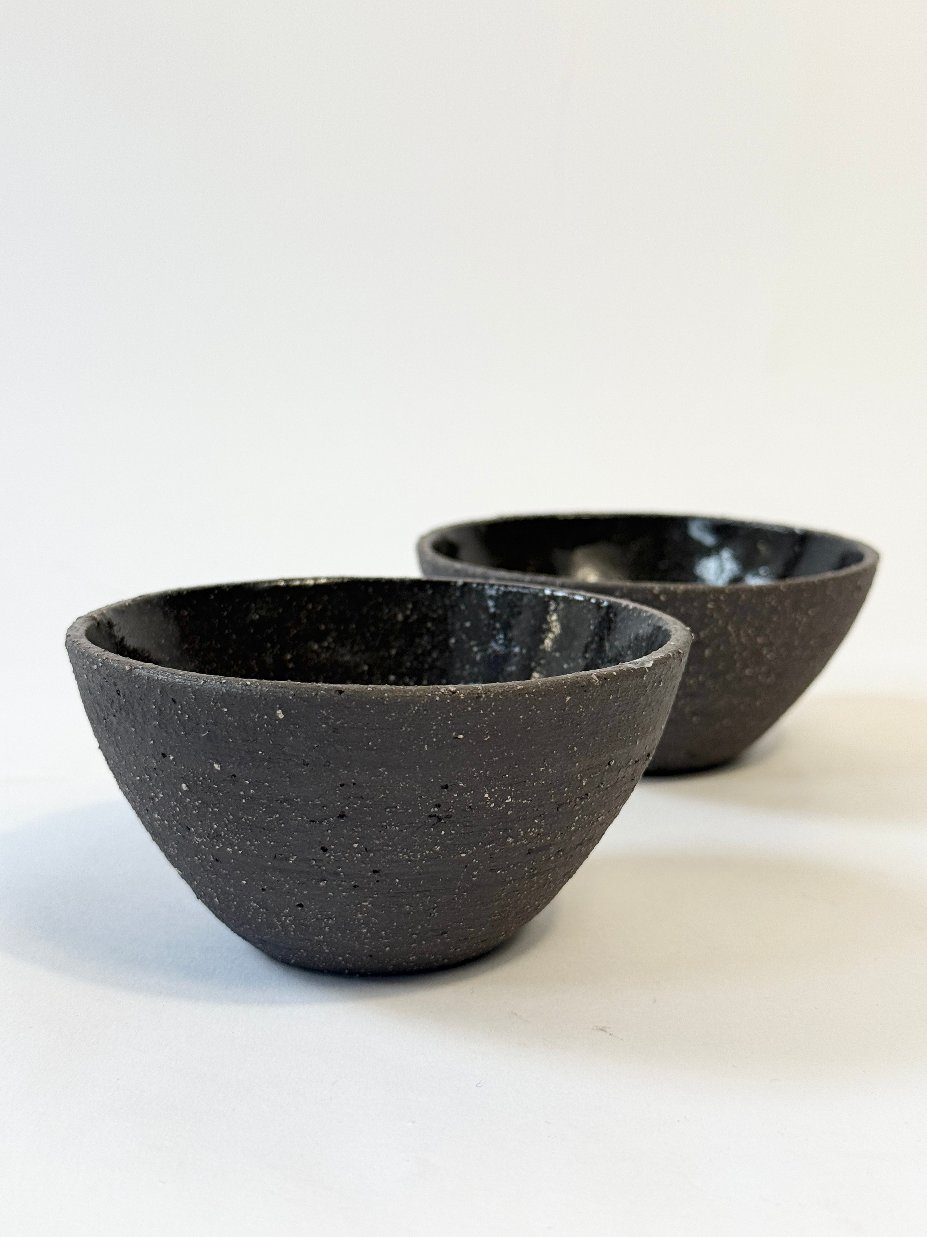 Two small handmade ceramic bowls with a textured, speckled dark matter exterior and a glossy black interior